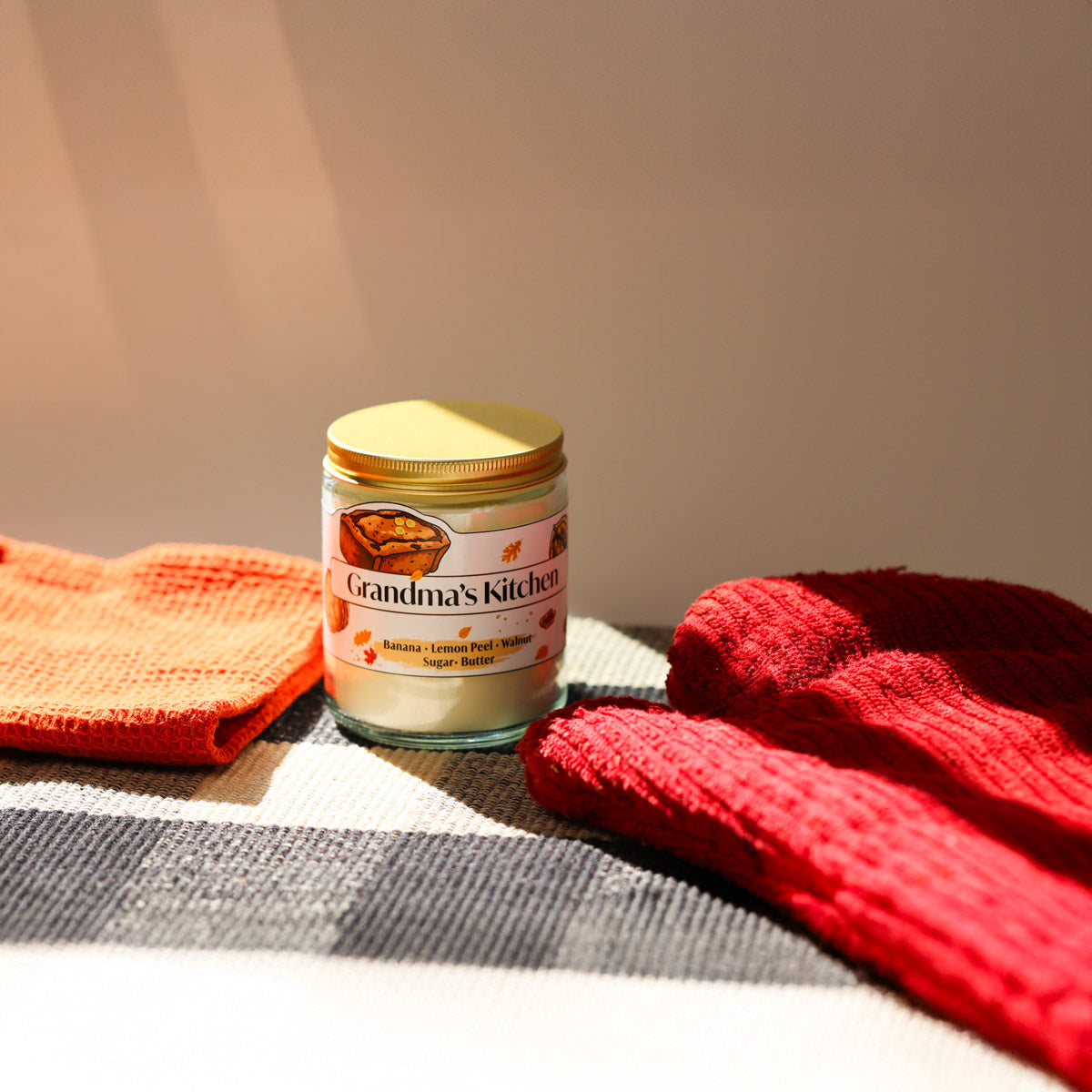 Jar of grandma's kitchen candle with red mitten and a kitchen towel on a checkered cloth.