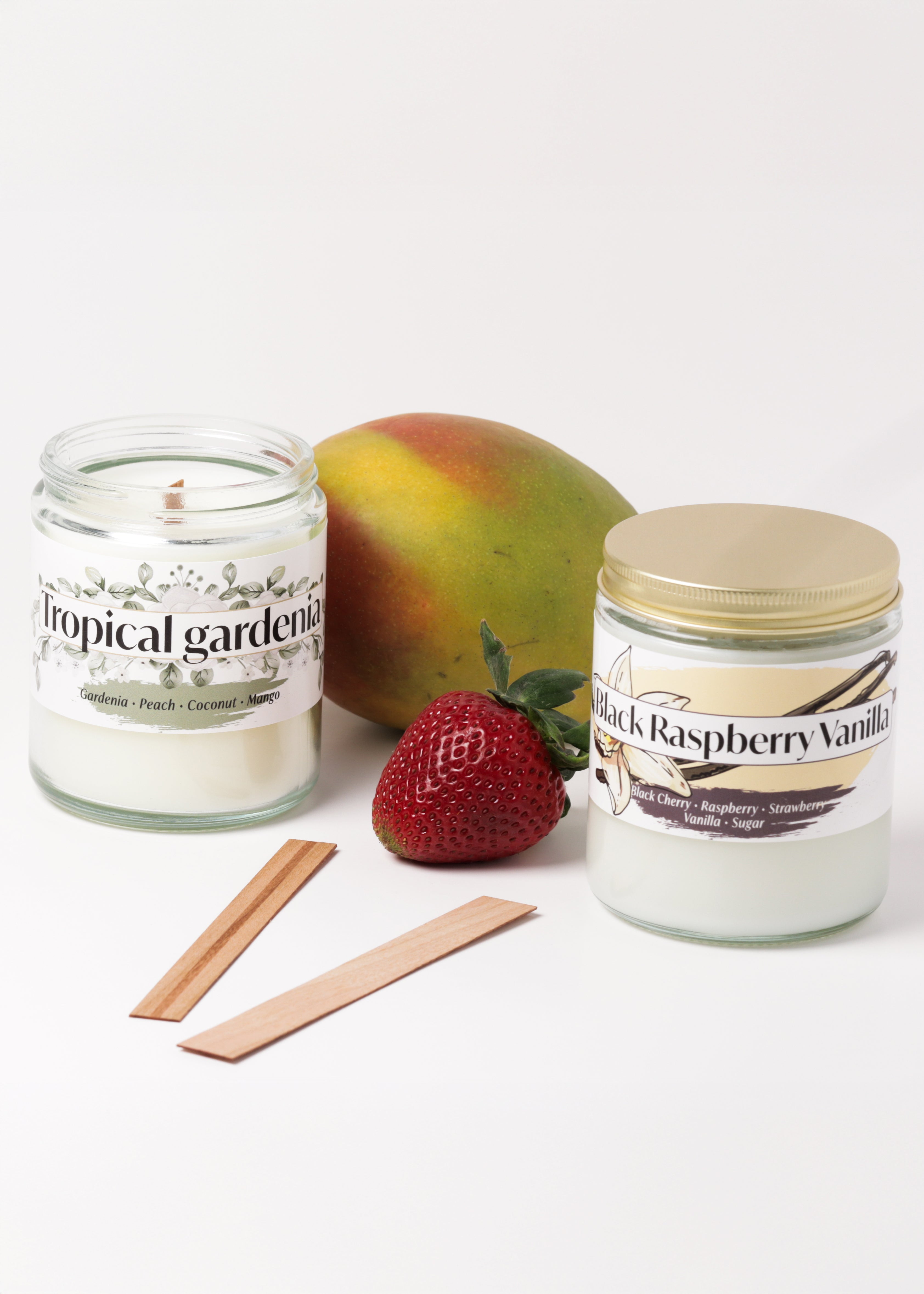 Two scented candles in glass jars with a mango, strawberry and two wooden wicks between them
