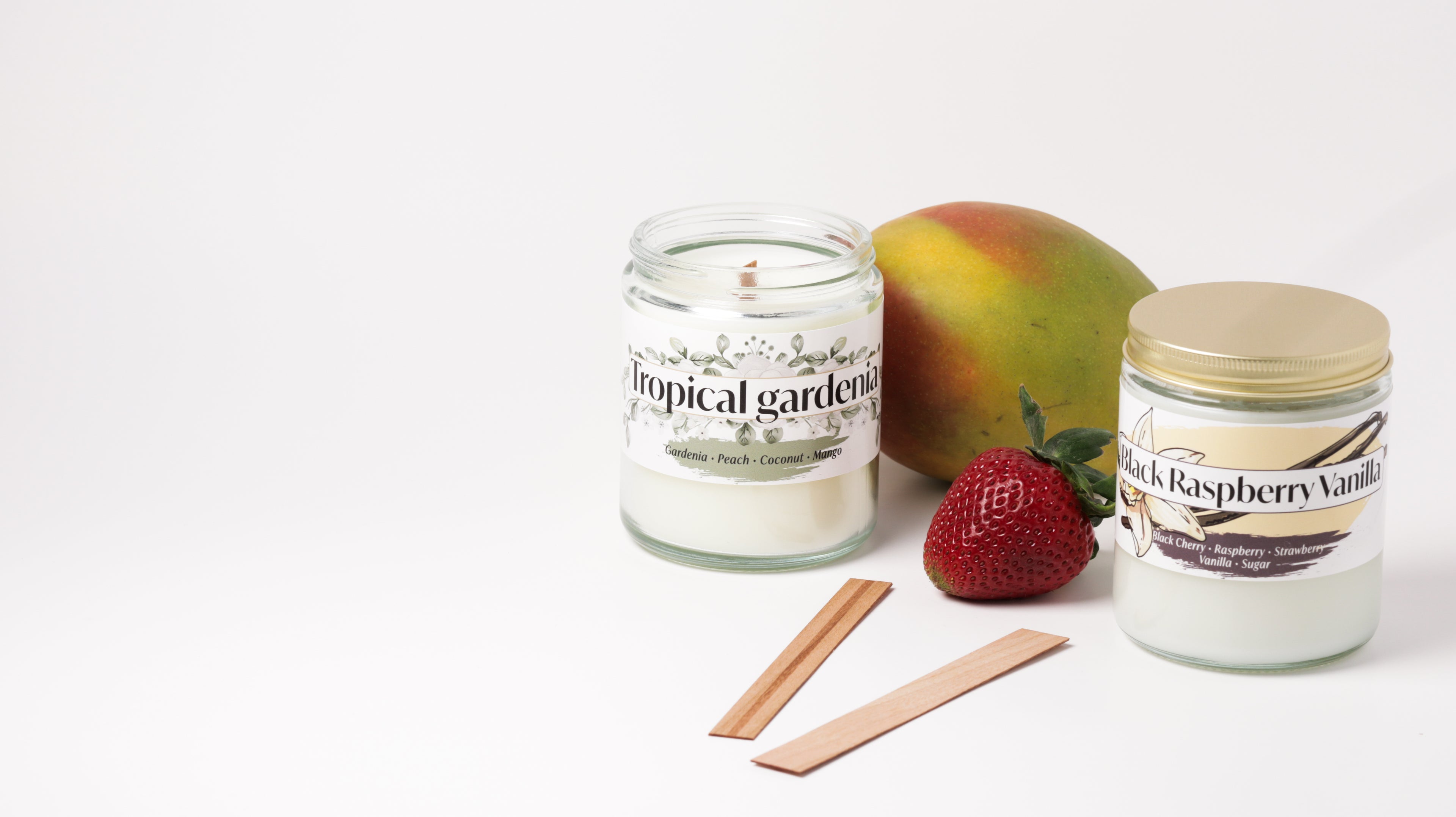 Two scented candles in glass jars with a mango, strawberry and two wooden wicks  between them